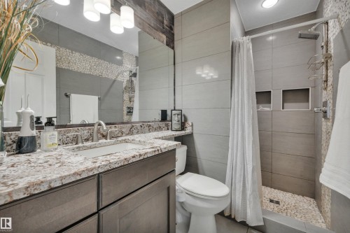 The bathroom features a vanity with a granite countertop and a white sink, a shower with grey tile walls and a pebble floor, and a toilet - 8807 31 Avenue, Edmonton, AB - Indoor Photo Showing Bathroom