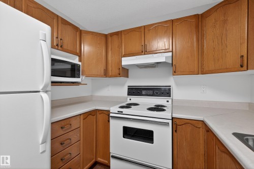 The kitchen features wood cabinetry, white countertops, and white appliances including a refrigerator, microwave, and range - 106 15499 Castle Downs Road, Edmonton, AB - Indoor Photo Showing Kitchen
