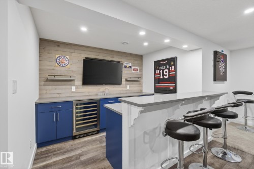 The wet bar features blue cabinetry, a stainless steel beverage refrigerator, a light-colored countertop, and a wood plank accent wall - 13 Bilodeau Crescent, Beaumont, AB - Indoor Photo Showing Other Room