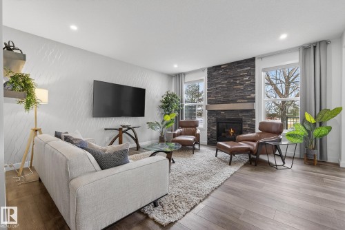 Living room featuring hardwood flooring, a fireplace with a stacked stone facade, and large windows with privacy curtains - 13 Bilodeau Crescent, Beaumont, AB - Indoor Photo Showing Living Room With Fireplace