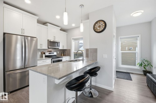 This kitchen features a central island with a light-colored countertop and seating, stainless steel appliances, white cabinetry, and light-toned flooring - 17498 76 St Nw, Edmonton, AB - Indoor Photo Showing Kitchen With Double Sink