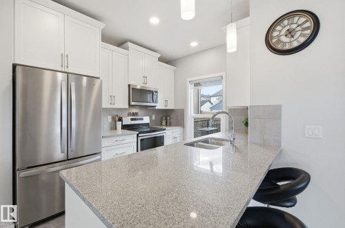 The kitchen features a stainless steel refrigerator and microwave, white cabinetry, a light-colored countertop, and a double basin sink with a chrome faucet - 17498 76 St Nw, Edmonton, AB - Indoor Photo Showing Kitchen With Double Sink With Upgraded Kitchen