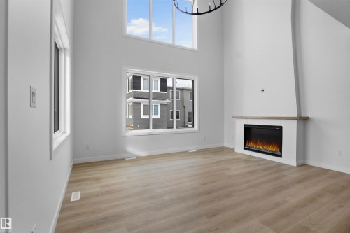 The property features a spacious living area with light wood flooring, a contemporary fireplace with a mantel, and high ceilings with tall windows - 3304 Chickadee Drive, Edmonton, AB - Indoor Photo Showing Other Room With Fireplace