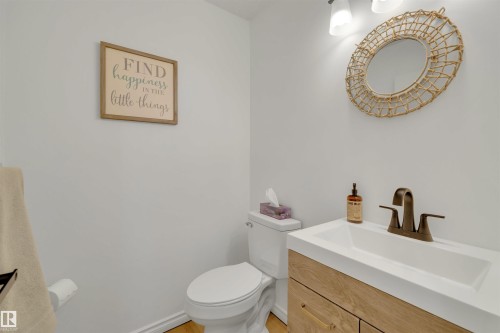 The bathroom features a toilet, a vanity with a white sink and bronze-toned faucet, and a round mirror with a woven frame - 131 West Liberty Crescent, Millet, AB - Indoor Photo Showing Bathroom