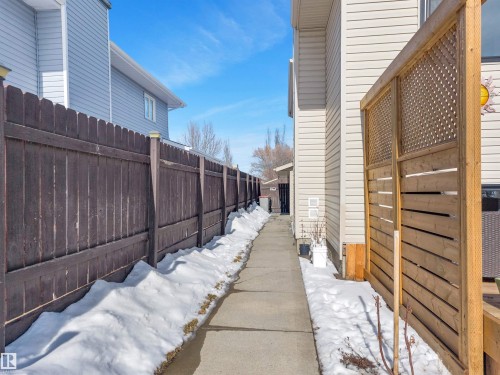 The property features a concrete pathway running alongside the exterior, bordered by a dark wooden fence and a light-colored privacy screen - 131 West Liberty Crescent, Millet, AB - Outdoor With Exterior