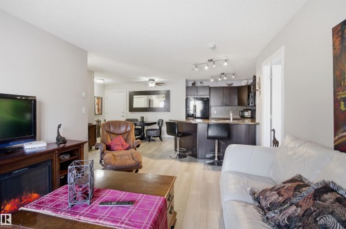 Open concept living area featuring light-colored flooring, a modern electric fireplace with a dark wood mantel, and a kitchen with dark cabinetry, black appliances, and track lighting - 115 2208 44 Avenue, Edmonton, AB - Indoor Photo Showing Living Room