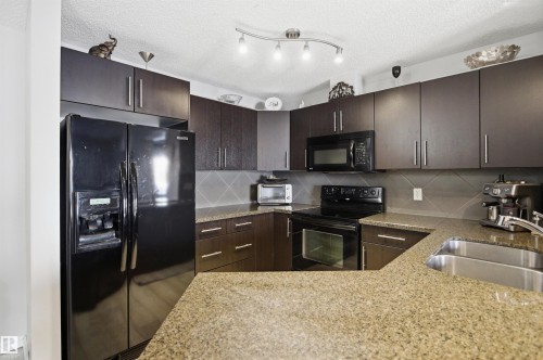 The kitchen features dark wood cabinetry, granite countertops, and black appliances including a refrigerator, microwave, and oven with stovetop - 115 2208 44 Avenue, Edmonton, AB - Indoor Photo Showing Kitchen With Double Sink