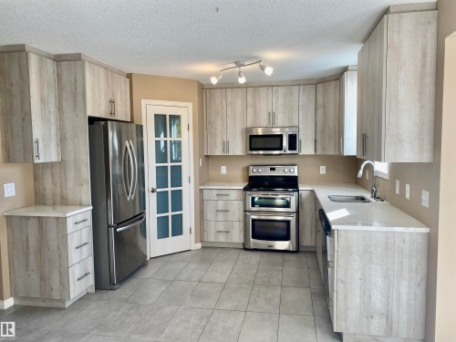 Kitchen with light wood finish cabinetry, stainless steel appliances, a textured ceiling, light stone counters, and light tile patterned floors - 403 178 Bridgeport Boulevard, Leduc, AB - Indoor Photo Showing Kitchen With Stainless Steel Kitchen