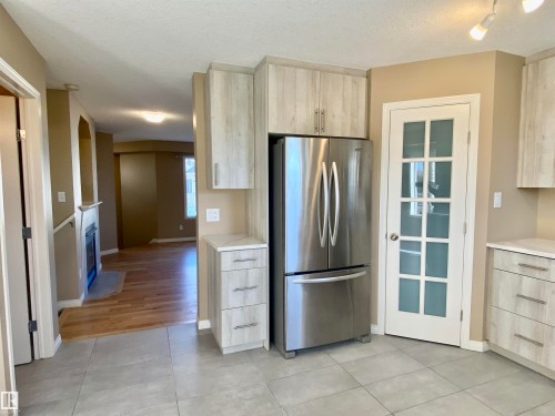 Kitchen featuring freestanding refrigerator, light wood finish cabinets, light tile patterned flooring, a textured ceiling, and a fireplace - 403 178 Bridgeport Boulevard, Leduc, AB - Indoor Photo Showing Kitchen With Stainless Steel Kitchen