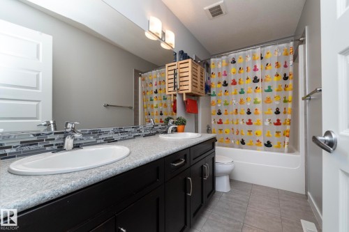 Bathroom featuring a vanity with two sinks, dark cabinetry, and a textured countertop, complemented by a tiled backsplash - 7608 179 Avenue, Edmonton, AB - Indoor Photo Showing Bathroom