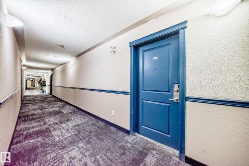 The property features a blue entry door with a contemporary door handle and a peephole, set within a hallway adorned with wall sconces and patterned carpeting - 126 16221 95 Street, Edmonton, AB - Indoor Photo Showing Other Room