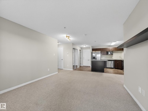 This property features a spacious open-concept living area with carpeting, leading into a kitchen equipped with stainless steel appliances, dark cabinetry, and an island - 1111 9363 Simpson Drive, Edmonton, AB - Indoor