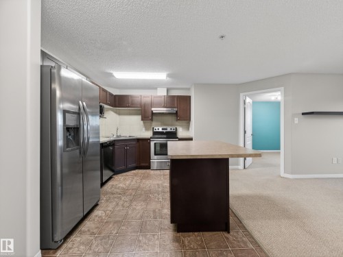 The kitchen features a stainless steel side-by-side refrigerator, a stainless steel oven and range, dark wood cabinetry, and a central island with a light-colored countertop - 1111 9363 Simpson Drive, Edmonton, AB - Indoor Photo Showing Kitchen