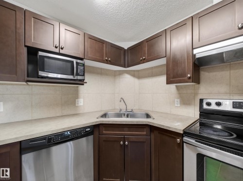 The kitchen features dark wood cabinetry, a stainless steel microwave, a stainless steel dishwasher, a stainless steel sink with a chrome faucet, and a black and stainless steel electric range - 1111 9363 Simpson Drive, Edmonton, AB - Indoor Photo Showing Kitchen With Double Sink