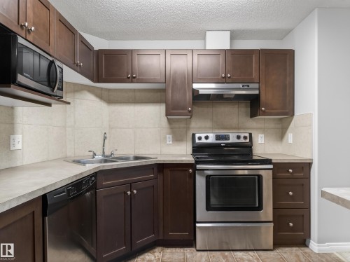 The kitchen features dark wood cabinetry, a stainless steel microwave, a double basin sink, stainless steel dishwasher, and a stainless steel range with an overhead range hood - 1111 9363 Simpson Drive, Edmonton, AB - Indoor Photo Showing Kitchen With Double Sink