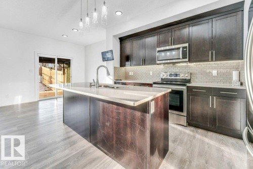 The kitchen features dark wood cabinetry, stainless steel appliances, a grey tiled backsplash, and a kitchen island with a sink and light-colored countertop - 1912 27 Street Nw, Edmonton, AB - Indoor Photo Showing Kitchen With Stainless Steel Kitchen With Upgraded Kitchen