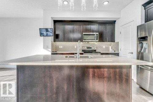 Kitchen featuring a central island with a light-colored countertop and an integrated sink, dark wood cabinetry, stainless steel appliances including a microwave and refrigerator, and a tiled backsplash - 1912 27 Street Nw, Edmonton, AB - Indoor Photo Showing Kitchen With Stainless Steel Kitchen With Upgraded Kitchen