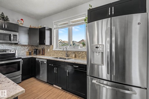 Kitchen featuring dark cabinetry, stainless steel appliances, light stone counters, a textured ceiling, and light wood-style floors - 4110 Allan Crescent, Edmonton, AB - Indoor Photo Showing Kitchen With Double Sink With Upgraded Kitchen