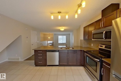 The kitchen features an island with a sink and dishwasher, dark wood cabinetry, stainless steel appliances including a refrigerator, microwave, and oven, and light-colored tiled flooring - 87 4050 Savaryn Drive, Edmonton, AB - Indoor Photo Showing Kitchen With Double Sink
