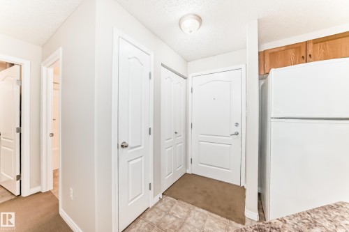 Entryway featuring an overhead light fixture, white doors with silver hardware, and a white refrigerator with wooden cabinetry above - 102 111 Edwards Drive, Edmonton, AB - Indoor Photo Showing Other Room