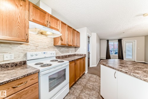 The kitchen features wood cabinetry, a white stove, and a complementary tile backsplash, with a peninsula providing additional counter space - 102 111 Edwards Drive, Edmonton, AB - Indoor Photo Showing Kitchen