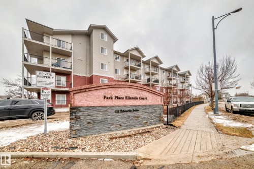The property features a multi-story building with balconies, a decorative stone and stucco sign, and a paved sidewalk - 102 111 Edwards Drive, Edmonton, AB - Outdoor