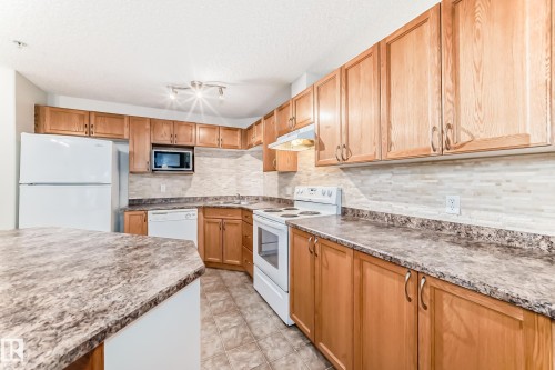 The kitchen features wood cabinetry, a tiled backsplash, and stone-look countertops - 102 111 Edwards Drive, Edmonton, AB - Indoor Photo Showing Kitchen
