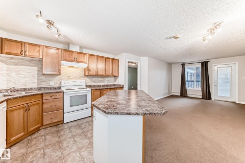 Open concept living area featuring a kitchen with wood cabinetry, tiled backsplash, and an island with a countertop, leading into a carpeted living space with a large window and an exterior door - 102 111 Edwards Drive, Edmonton, AB - Indoor Photo Showing Kitchen
