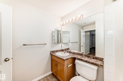 The bathroom features a vanity with a light wood base and a patterned countertop, a built-in mirror with overhead lighting, and a white toilet - 102 111 Edwards Drive, Edmonton, AB - Indoor Photo Showing Bathroom