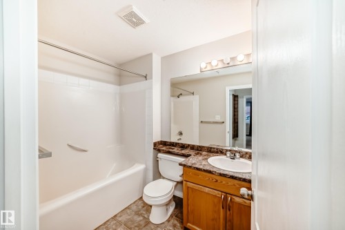The bathroom features a combination tub and shower with white tile surround, a vanity with a large mirror and overhead lighting, and a toilet - 102 111 Edwards Drive, Edmonton, AB - Indoor Photo Showing Bathroom
