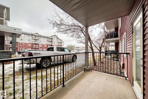 This property features a private covered patio with a concrete surface and black metal railings, offering views of the surrounding grounds and buildings - 102 111 Edwards Drive, Edmonton, AB - Outdoor With Exterior