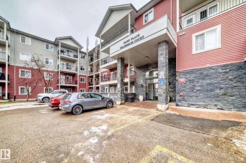 The property features a multi-story building with a red and grey exterior, balconies, and a stone facade entrance - 102 111 Edwards Drive, Edmonton, AB - Outdoor