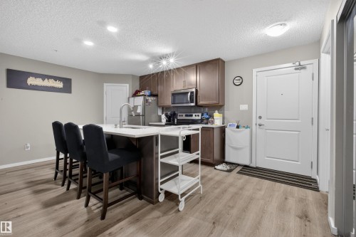 The property features an open-concept kitchen with wood cabinetry, stainless steel appliances, and a central island with an integrated sink and breakfast bar seating for three - 134 148 Ebbers Boulevard, Edmonton, AB - Indoor