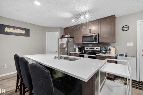 The kitchen features a spacious island with a white countertop and seating for three, stainless steel appliances, and wood cabinetry - 134 148 Ebbers Boulevard, Edmonton, AB - Indoor Photo Showing Kitchen With Double Sink