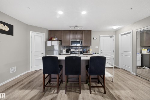 This kitchen features an island with a light-colored countertop and seating, stainless steel appliances, dark wood cabinetry, and light-toned flooring - 134 148 Ebbers Boulevard, Edmonton, AB - Indoor Photo Showing Kitchen