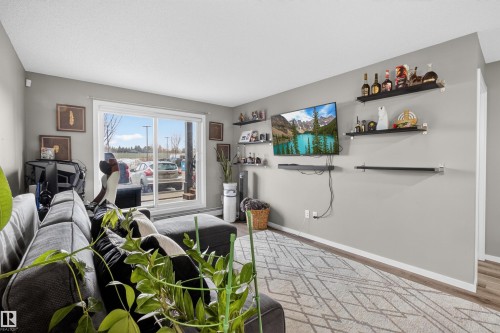The living room features light gray walls, a sliding glass door, and a television mounted on the wall - 134 148 Ebbers Boulevard, Edmonton, AB - Indoor