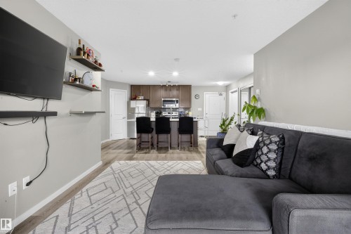 Open concept living area featuring light gray walls, wood-style flooring, and a kitchen with dark cabinetry and stainless steel appliances - 134 148 Ebbers Boulevard, Edmonton, AB - Indoor Photo Showing Living Room