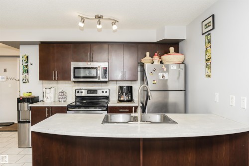 The kitchen features dark wood cabinetry, a stainless steel sink with a high arc faucet, and stainless steel appliances including a microwave, refrigerator, and range - 107 10118 106 Avenue, Edmonton, AB - Indoor Photo Showing Kitchen With Double Sink