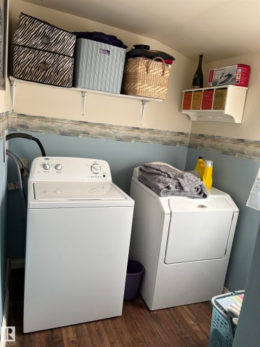 This functional laundry area features a washer and dryer, wood-look flooring, and a wall-mounted shelving unit for storage - 5117 52 Avenue, Elk Point, AB - Indoor Photo Showing Laundry Room