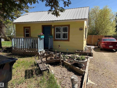 The property features yellow siding, a metal roof, and a front porch with a blue railing. Fenced back yard with parking spaces behind. Beautiful flower garden and landscaped front yard. - 5117 52 Avenue, Elk Point, AB - Outdoor