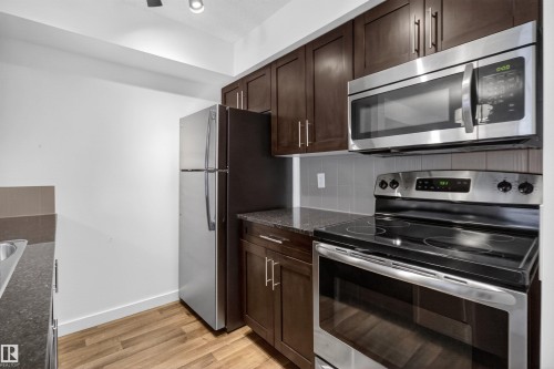 The kitchen features dark wood cabinetry, stainless steel appliances, a grey tiled backsplash, and light wood flooring - 311 12035 22 Avenue, Edmonton, AB - Indoor Photo Showing Kitchen