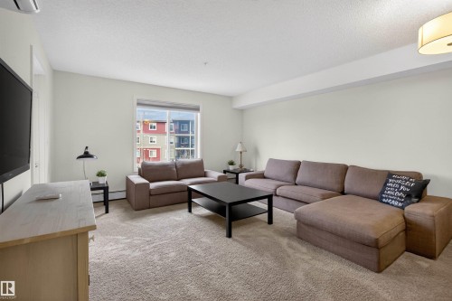 The living area features light-colored walls, a large window providing natural light, and a light-colored carpet - 311 12035 22 Avenue, Edmonton, AB - Indoor Photo Showing Living Room