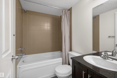 Bathroom featuring a white bathtub with a shower, brown tiled walls, a white toilet, and a vanity with a dark countertop and an integrated sink - 311 12035 22 Avenue, Edmonton, AB - Indoor Photo Showing Bathroom
