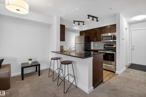 The kitchen features stainless steel appliances, dark wood cabinetry, and a black countertop with an integrated sink - 311 12035 22 Avenue, Edmonton, AB - Indoor Photo Showing Kitchen With Stainless Steel Kitchen