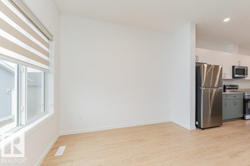 This bright room features light-colored flooring, a large window with striped blinds, and a view of the adjacent kitchen with stainless steel appliances - 9652 Colak Link Link, Edmonton, AB - Indoor