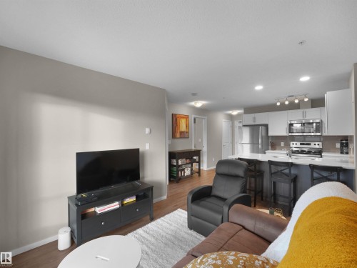 The open concept living area features hardwood flooring, recessed lighting, and a kitchen with stainless steel appliances, white cabinetry, and an island with seating - 214 394 Windermere Road, Edmonton, AB - Indoor Photo Showing Living Room