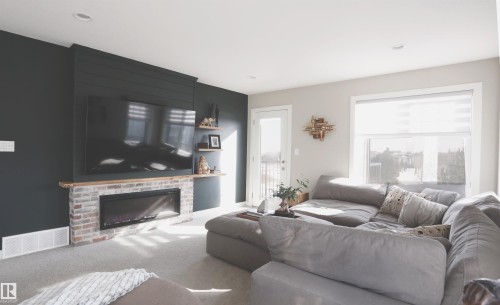 The living area features a dark accent wall with a fireplace, light-colored carpeting, a window with roller blinds, and a door leading to an outdoor space - 1003 Huckell Place, Edmonton, AB - Indoor Photo Showing Living Room With Fireplace
