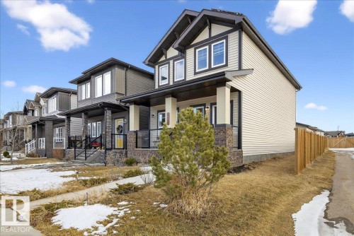The property presents a modern exterior with light-colored siding, dark trim, and stone accents on the front porch pillars - 203 Cavanagh Common, Edmonton, AB - Outdoor With Deck Patio Veranda With Facade