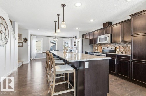 The kitchen features a large island with seating, dark wood cabinetry, stainless steel appliances, and a tiled backsplash - 16720 15 Avenue, Edmonton, AB - Indoor Photo Showing Kitchen With Upgraded Kitchen