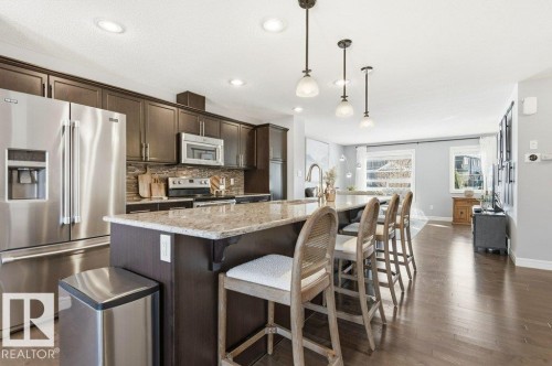 The kitchen features dark wood cabinetry, stainless steel appliances, a tiled backsplash, and an island with a light-colored countertop and seating - 16720 15 Avenue, Edmonton, AB - Indoor Photo Showing Kitchen With Upgraded Kitchen